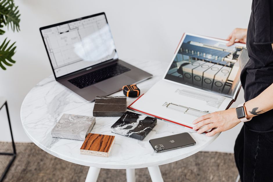 Architect reviews design plans on a marble table with laptop, phone, and material samples.